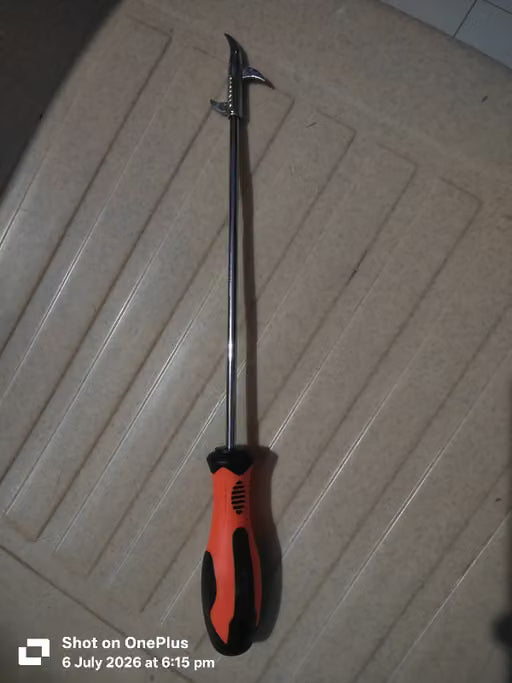 A long-handled metal tool with a curved silver tip and an orange-and-black handle, photographed on a beige textured floor. A OnePlus timestamp watermark is visible at the bottom left.