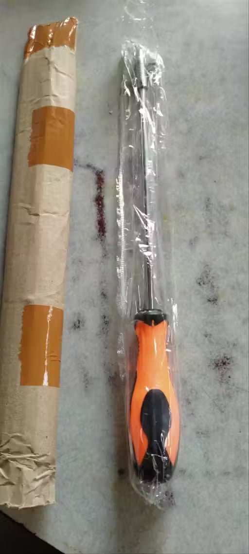 An orange and black screwdriver wrapped in clear plastic packaging, placed next to its cardboard tube with orange tape on a gray marble-like surface.