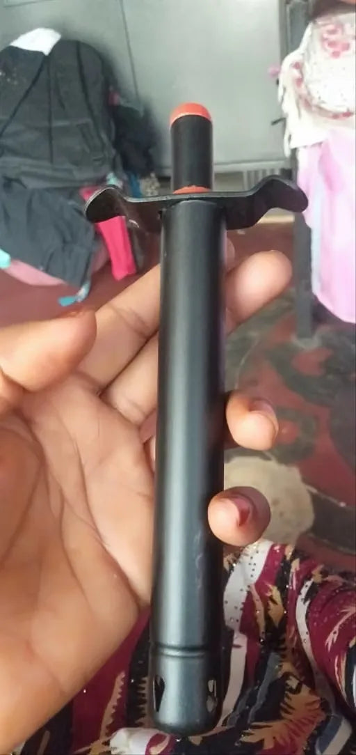 A hand holding a black cylindrical tool or accessory with a small orange tip and a curved black tab on one end, photographed in a casual indoor setting.