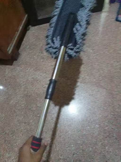 A long-handled microfiber duster mop being held by a hand on a speckled brown floor, with a wooden cabinet visible in the background.
