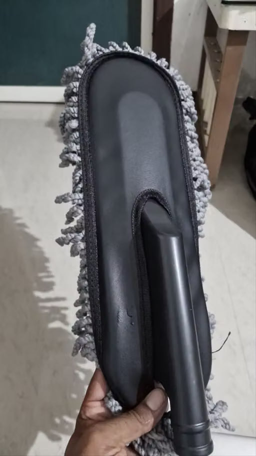 A person's hand holding a black plastic microfiber duster mop head with gray cleaning fibers, shown against an indoor tiled floor background.