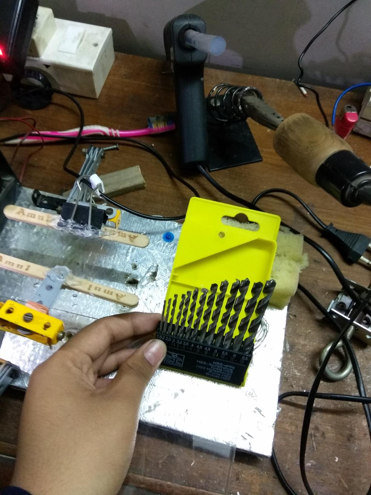Hand holding a set of drill bits in front of a workbench with tools and equipment.