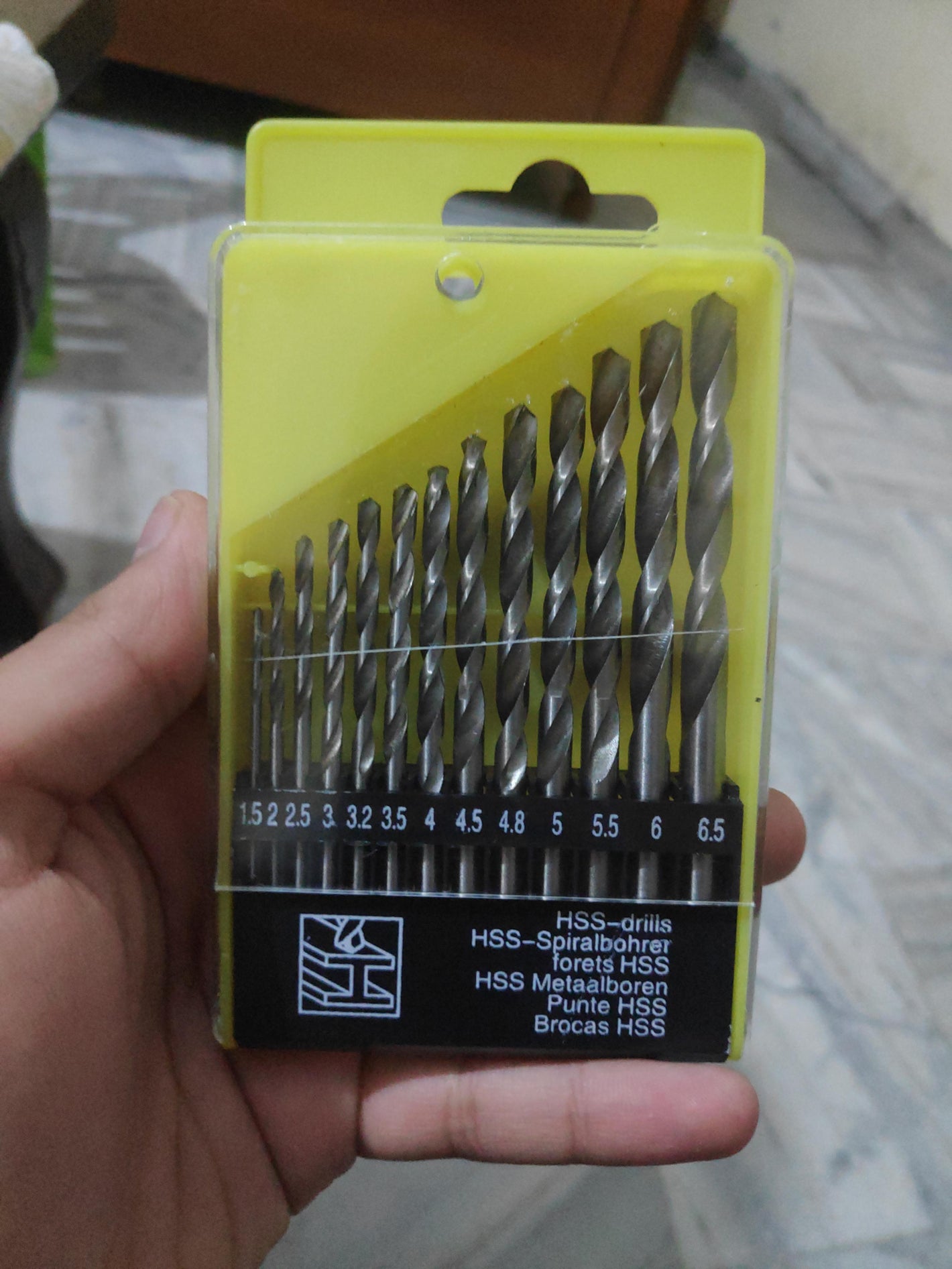 Set of drill bits in a yellow packaging held by a hand on a patterned surface.