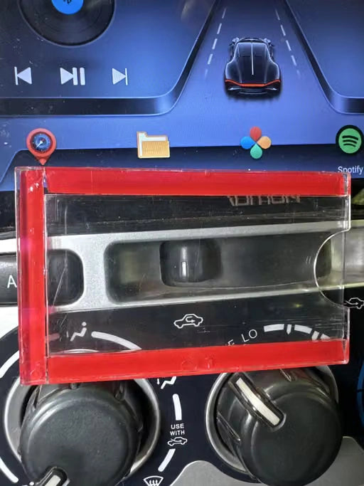A clear plastic cassette-style car dashboard accessory with a red frame, shown in front of a car infotainment screen and climate controls.