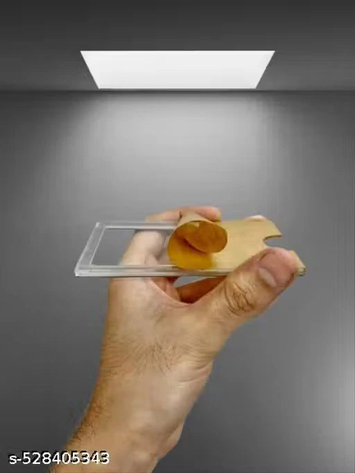A hand holds a clear rectangular plastic slide with a small amber-brown circular object inside, against a gray studio background with a bright light source above.