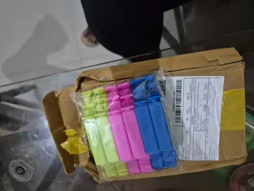 A brown cardboard box with a shipping label, containing several small wrapped items in bright neon green, pink, purple, and blue, photographed on a glass table.