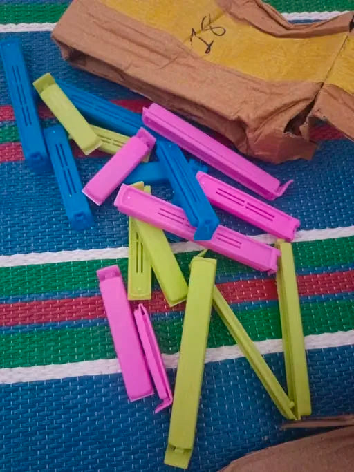 A pile of colorful plastic zipper clip bags in pink, blue, and green, scattered on a striped blue, green, and red woven mat with a brown paper bag in the background.