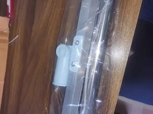 A close-up of a white plastic cabinet door pull handle attached to a metal track, wrapped in clear protective plastic on a wooden surface.