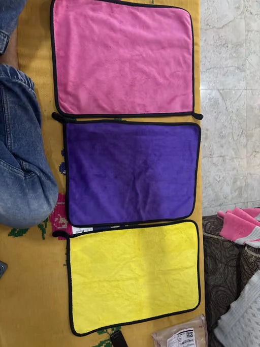 Three rectangular microfiber cloths in pink, purple, and yellow with black borders, laid out vertically on a yellow surface.