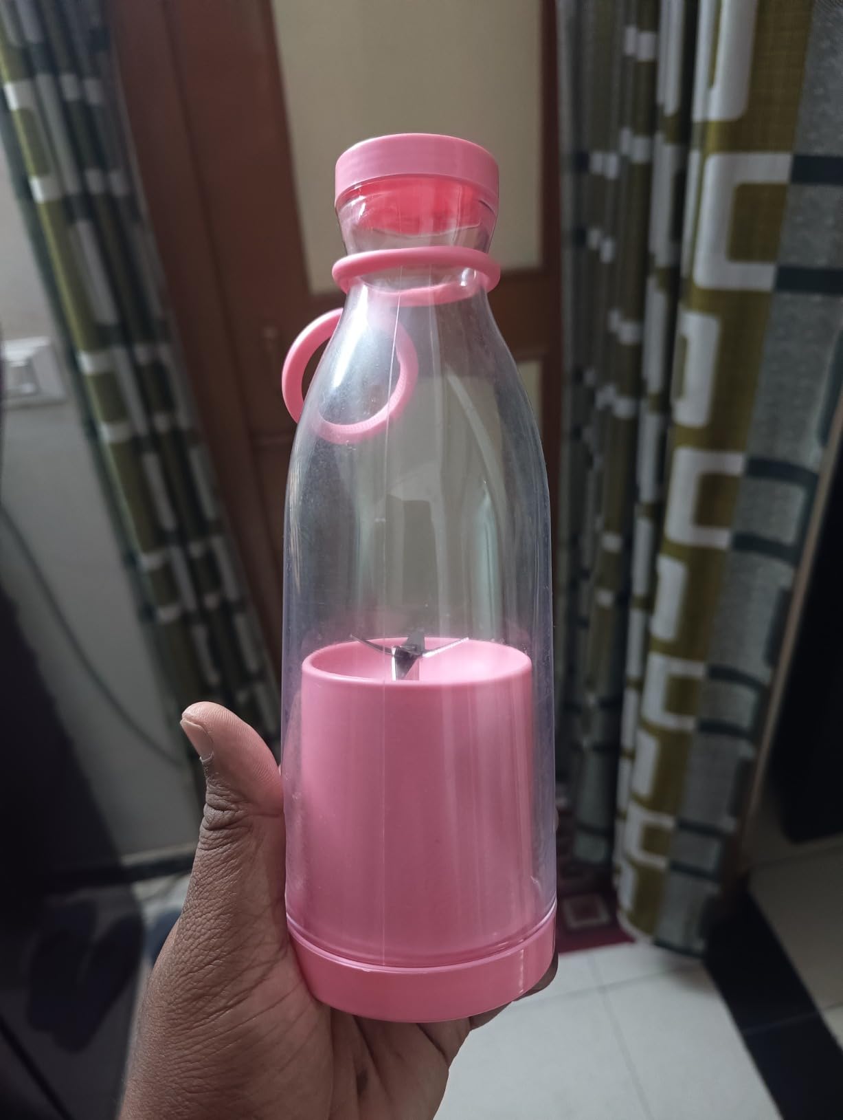 Pink water bottle with a transparent section held by a hand against a patterned curtain background.