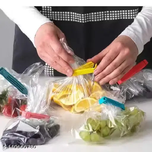 A person wearing a white shirt and black apron demonstrates using colorful plastic bag clips to seal clear bags containing sliced oranges, grapes, strawberries, and dark berries on a white countertop.