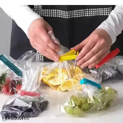 A person wearing a white shirt and black apron demonstrates using colorful plastic bag clips to seal clear bags containing sliced oranges, grapes, strawberries, and dark berries on a white countertop.