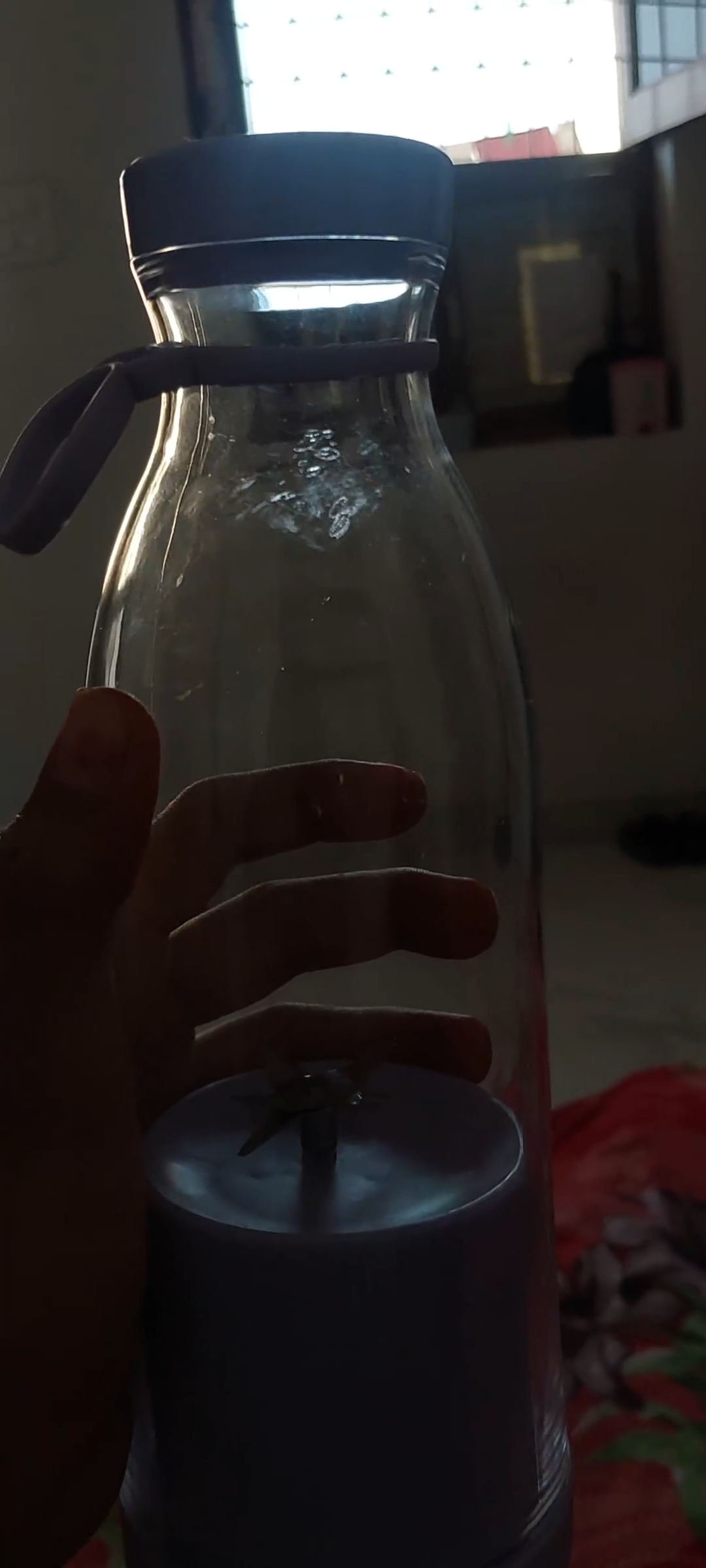 A hand holding a clear glass bottle with a dark gray lid and a matching dark gray base, shown in a dim indoor setting.