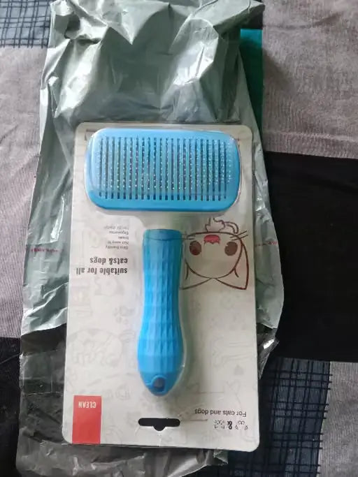A blue pet grooming brush in its original retail packaging, placed on top of a crumpled silver shipping mailer on a patterned fabric surface.