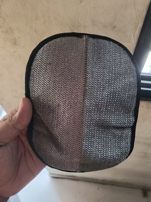 A hand holding a round, oval-shaped gray mesh pad with a black fabric border, set against a background of a beige wall and a window.