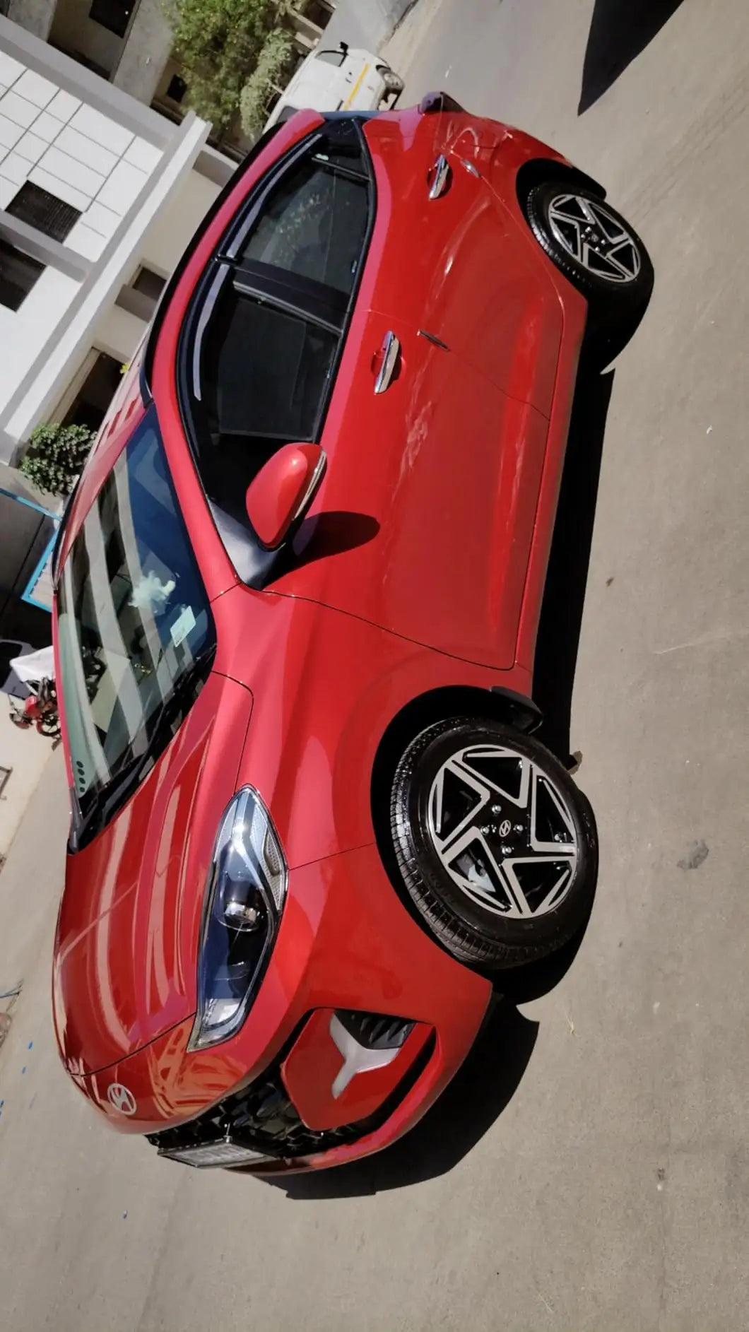 A red compact hatchback car photographed from a high-angle outdoor street setting, showing the front, side, and rear quarter panels with visible wheel and shadow details.