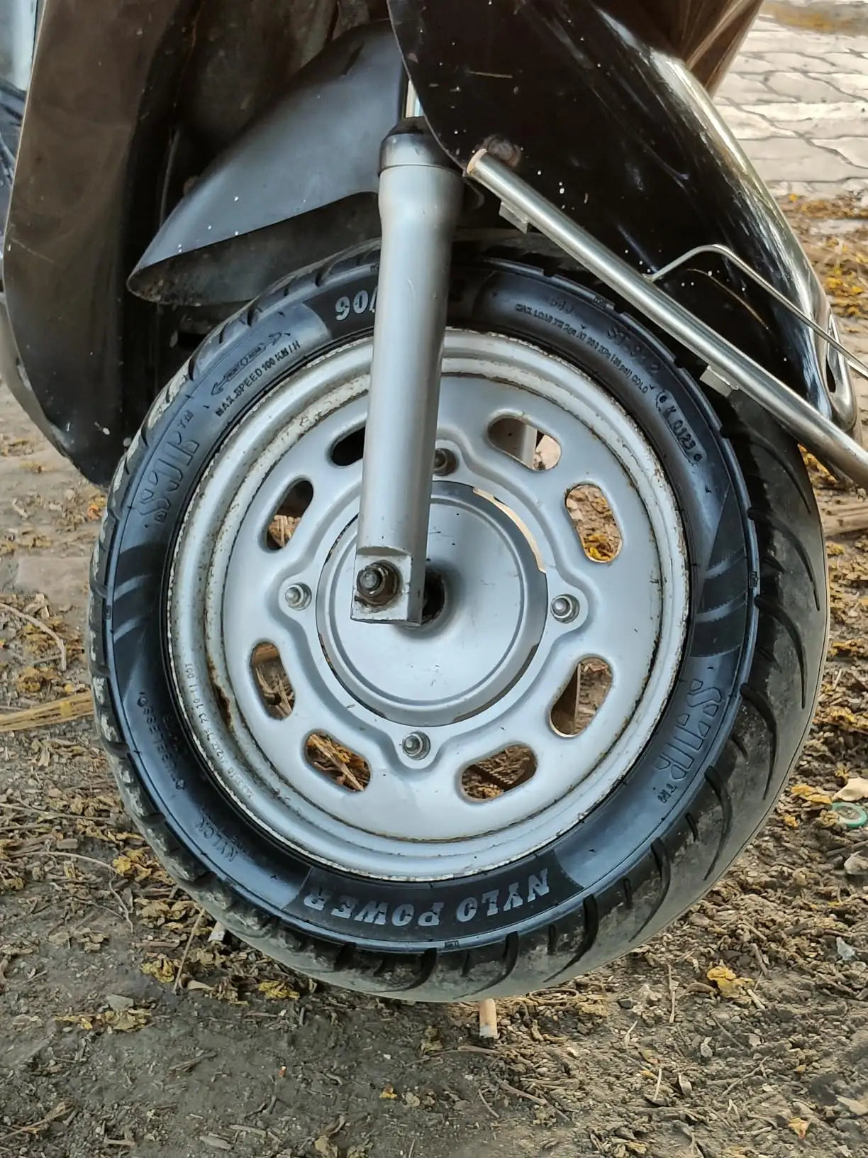 Close-up of a scooter's front wheel with a silver metal rim, black tire, and visible suspension fork, photographed outdoors on dirt ground.