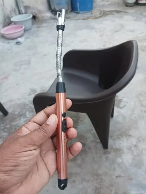 A hand holding a flexible metal electric lighter with a metallic rose gold handle and black accents, with a dark brown plastic chair in the background on a concrete floor.